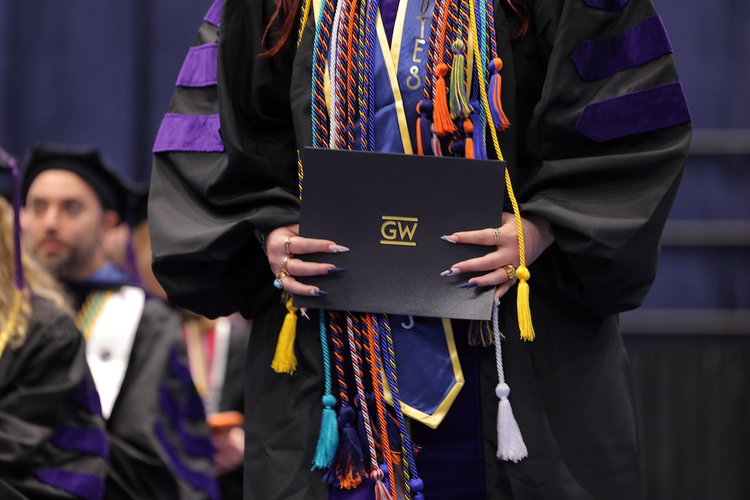 A woman in a graduation gown with chords holding a GW diploma during graduation.