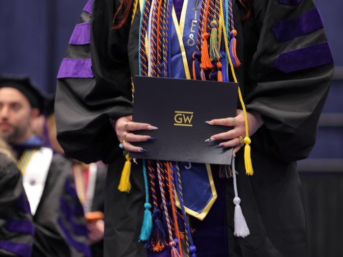 A woman in a graduation gown with chords holding a GW diploma during graduation.