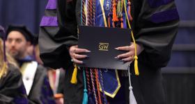 A woman in a graduation gown with chords holding a GW diploma during graduation.